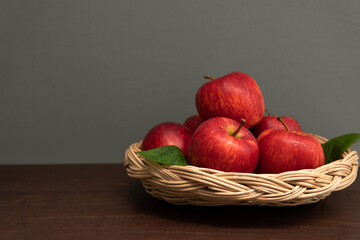 Still life fresh heap of red apple with leaves in a wicker basket on dark brown wooden table against grey color wall background with copy space