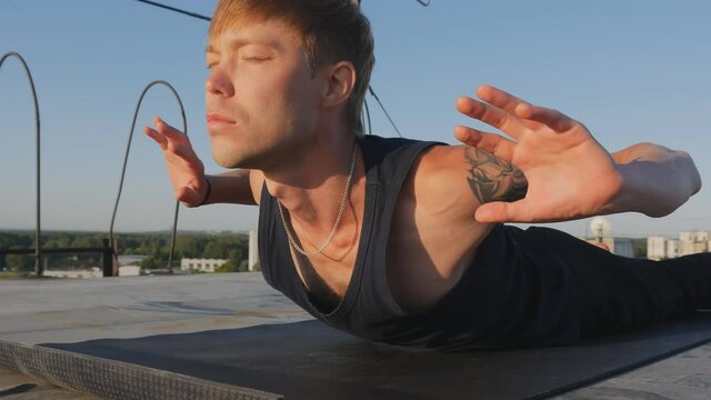 a handsome man with a tattoo and a beautiful body doing yoga on the rooftop in the morning sunlight. on the mat, lying on his stomach, doing the assan arching his back. Wide angle lens