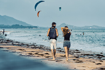 Young couple walks along the sandy beach at sunset in summer. Kite-surfings are riding in the background.