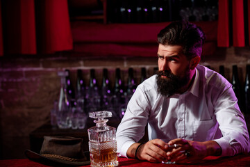 Handsome pensive man is holding a glass of whiskey and looking away while sitting in bar indoors.