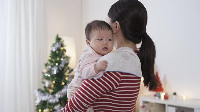 asian new mother standing with cloth on shoulder in a bright Christmas living room is turning around while burping her baby at home.
