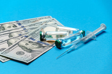 Close up 100 US dollar bills, syringe and vaccine ampoules over blue background