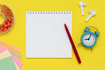 Notebook and pen on a yellow background. Notepad, white headphones, alarm clock, clips, and colorful stickers on workplace. Template with copy space, flat design, mockup. Planning concept, education.