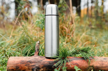 Thermos in a coniferous forest, close-up. Vacuum travel bottle stands on a tree stump. Concept for hiking, travel, hot drink in cold weather.