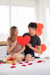 A young couple lovers hold a red heart shape and eye contact each other on their bedroom which have a red and a golden gift box on the bed.