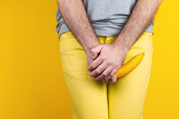 Potency and men's health. A man in yellow jeans holds a banana in his hands, confused, at the level of the genitals. Yellow background. Close up