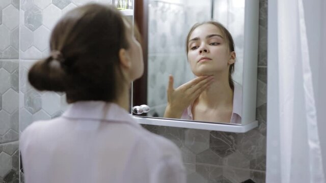 Charming woman with natural beautiful skin applying cosmetic daily cream on neck and decollete while standing in bathroom in morning and doing everyday skin care procedure in front of mirror 