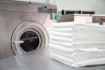 Stack of clean bed sheets in front of industrial washing machines. Focused on washing machines. 
Shot taken in the factory.