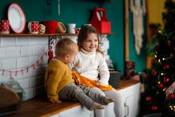 Toddler with older sister children hugging in the new year's kitchen, the concept of cozy christmas and new year, decorated house, lifestyle in a green red festive interior