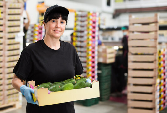 Portrait of woman warehouse worker carrying box with fresh avocado fruits on packing facility