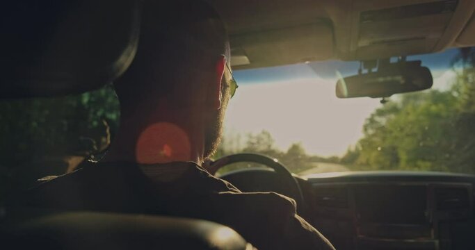 Man drives the car while sunset.  Young man driving the car.  Young adult boy driving car. Real time. Sun's rays shine through the windshield of the car. Rear View to the man drives a car.