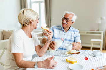 Fototapeta na wymiar Elderly couple sitting at the table. Senior man pouring milk into a cup. Married couple having breakfast at home. Mature woman holding peice of bread