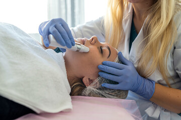 beautician in blue gloves makes a procedure of ultrasonic cleaning face to young female patient