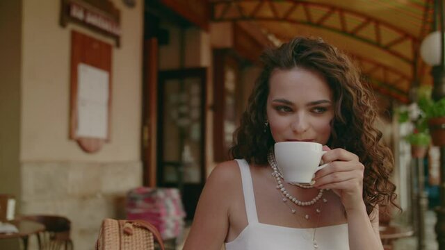 Curly brunette woman drinks coffee in street cafe in European city. Lifestyle conception. Close up portrait