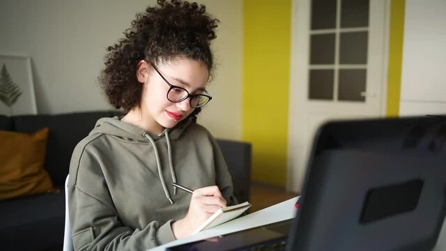mixed race teen student study online. girl looks at laptop and writes synopsis in notebook. concept e-learning, home school