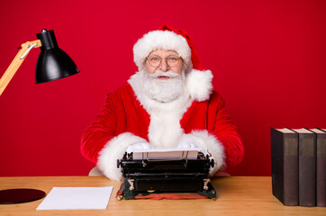 Photo of santa claus grey beard sit desk type vintage typewriter lamp paper book wear x-mas costume coat cap glasses isolated red color background