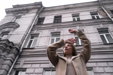 Obraz na płótnie Canvas a girl in a gray coat stands against the background of an architectural building, raising her hands to the top