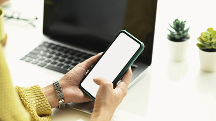 Cropped shot of a young female in yellow sweater hands holding smart phone with blank screen while sitting in front of laptop.