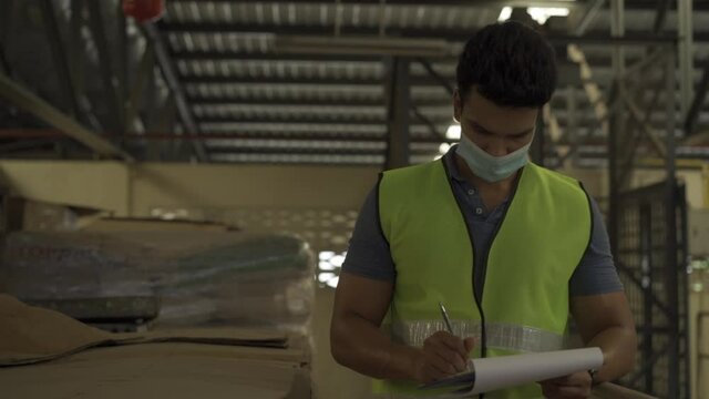 Young Indian factory warehouse worker wearing a protective face mask and safety helmet while working. 30s man checking stock order during Coronavirus Covid 19 pandemic - HD Slow Motion Footage