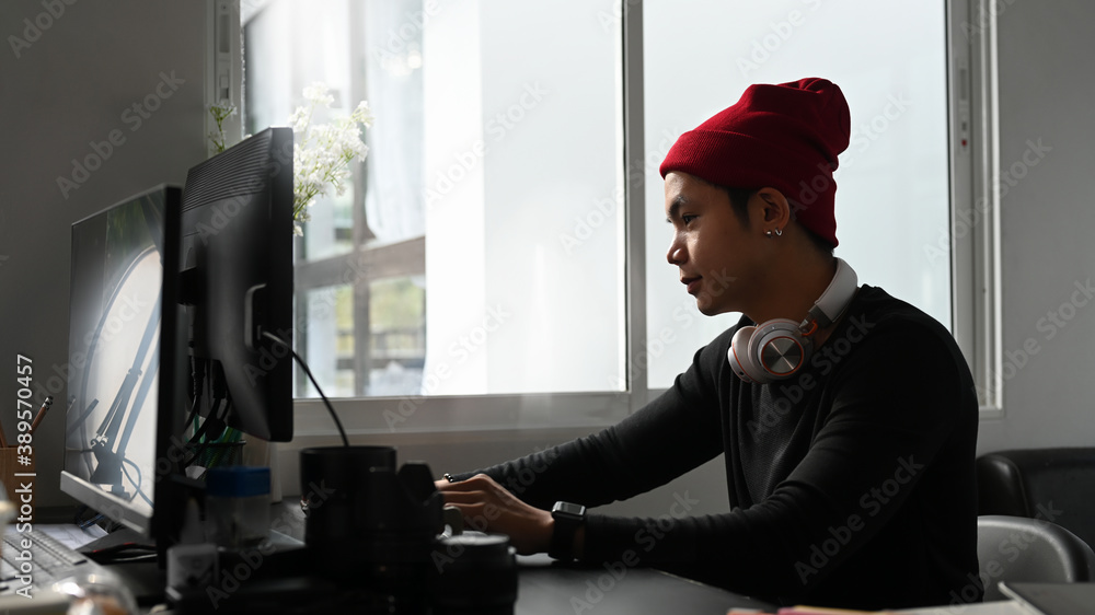 Aufkleber side view of young photographer in red wool hat working with new project on computer at his studio.