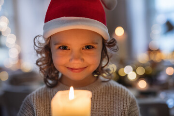 Cheerful small girl with Santa hat indoors at Christmas, holding candle.