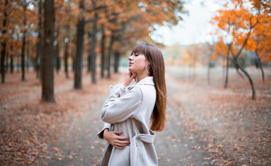 stylish young caucasian female with dark hair in the light coat on the alley in autumn park, trees...