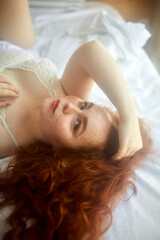 Obraz na płótnie Canvas lovely young woman with natural red hair posing at camera, lying on bed alone, coquette female in white lingerie, underwear, relax