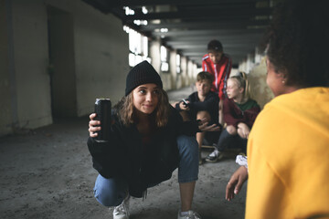 Group of teenagers gang sitting indoors in abandoned building, using smartphones.