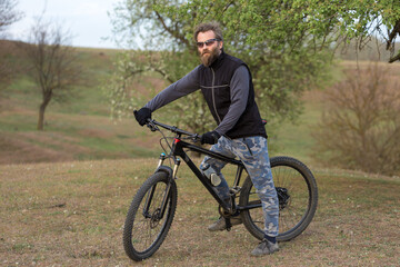 Sports brutal bearded guy on a modern mountain bike. Cyclist on the green hills in the spring.