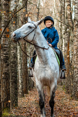 Little boy in a jockey cap on a white adult horse on a background of nature. Jockey, hippodrome,...
