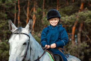 Little boy in a jockey cap on a white adult horse on a background of nature. Jockey, hippodrome,...