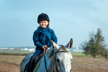 Little boy in a jockey cap on a white adult horse on a background of nature. Jockey, hippodrome,...