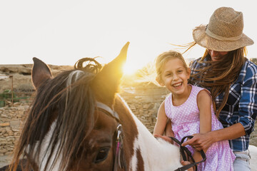 Happy family mother and daughter having fun riding horse inside ranch - obrazy, fototapety, plakaty
