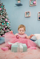 beautiful little girl in a tutu dress is sitting on the bed and looking at a gift on the background of a Christmas tree