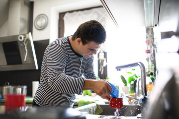 Down syndrome adult man standing indoors in kitchen at home, washing the dishes.