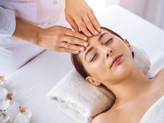 Beautiful brunette woman enjoying facial massage with closed eyes in sunny spa center . Relaxing treatment and cosmetic medicine concepts
