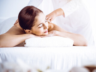 Fototapeta na wymiar Beautiful brunette woman enjoying back massage in sunny spa center. Beauty and medicine concept