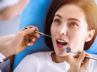 Smiling brunette woman being examined by dentist at sunny dental clinic. Hands of a doctor holding dental instruments near patient's mouth. Healthy teeth and medicine concept