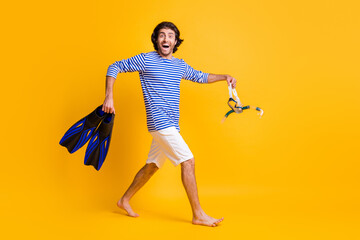 Full size profile side photo of guy hold scuba diving gear fins mask go wear white blue shorts isolated on yellow color background