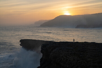 huge storm surge ocean waves crashing onto shore and cliffs at sunrise with a person standing there