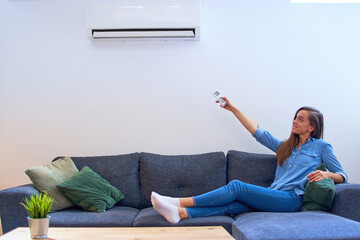 Young happy woman sitting on couch under air conditioner and adjusting comfort temperature with remote control at modern home