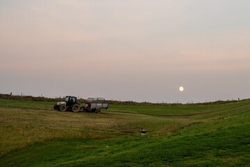 farmer with tractor and trailer cultivating fields under a full moon at sunset