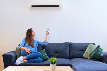 Young happy woman sitting on couch under air conditioner and adjusting comfort temperature with...