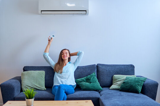 Young happy woman with closed eyes sitting on sofa under air conditioning and adjusting comfortable temperature with remote control at modern home