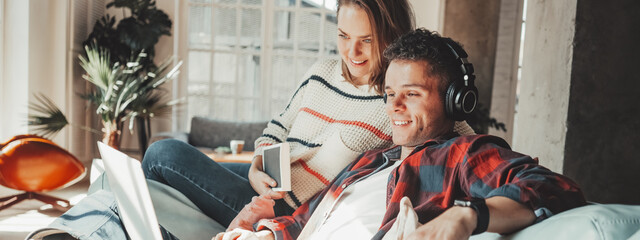 Fototapeta premium Young couple at home. Husband and wife sitting on the couch and watching TV series on laptop, listening music and reading books. Bright loft apartment