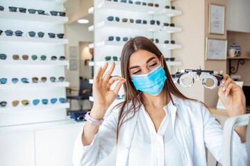 Beautiful young ophthalmologist in clinic with protective face mask. Portrait of smiling optometrist holding messbrille in ophthalmology clinic