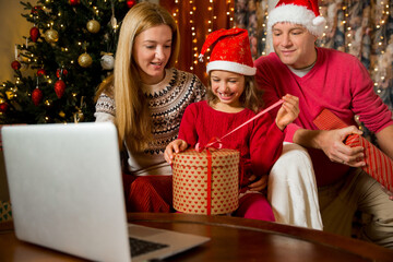 Fototapeta na wymiar A happy couple with a child is celebrating Christmas with their friends on video call using webcam. Family greeting their relatives on Christmas eve online. New normal