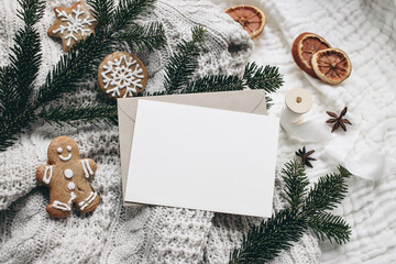 Christmas festive still life. Blank greeting card, invitation mockup and envelope on knitted sweater . Gingerbread cookies, dry oranges and fir branches on wool blanket. Selective focus, top view.