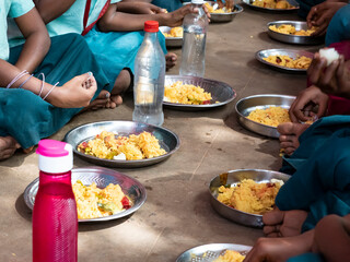 Unidentified poor classmates children with uniforms sitting on the floor outdoors, eating with their right hand some rice with masala. Lunch time