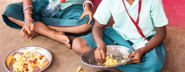 Unidentified poor classmates children with uniforms sitting on the floor outdoors, eating with...